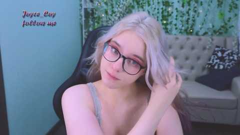 Media: Video of a young, slender, light-skinned woman with platinum blonde hair and large glasses, wearing a gray tank top, sitting in a black chair in a cozy, modern room with greenery and gray couch.