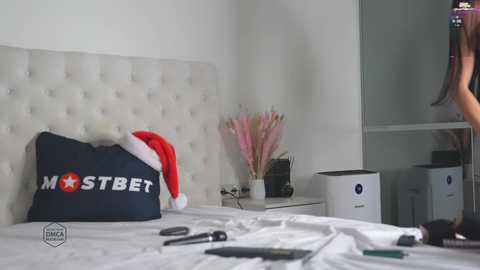 Media: Video of a modern bedroom with a white tufted headboard, a navy pillow with \"MOSTBET\" logo, a red Santa hat, a white air purifier, and scattered makeup brushes on a white bed.