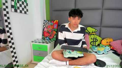 Media: Video of a young, dark-haired Asian boy in a striped polo, sitting cross-legged on a bed, holding a remote, surrounded by colorful toys and a green-and-white checkered wall.