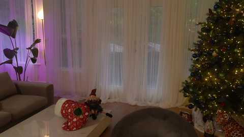 Media: A video of a cozy living room with a decorated Christmas tree, a plush toy, and sheer curtains, illuminated by soft purple and white light.