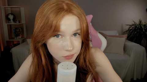 Media: Video of a fair-skinned, red-haired woman with blue eyes, wearing a pink top, holding a white Nintendo Switch controller. She is in a cozy, dimly lit room with a beige couch and pink pillows.