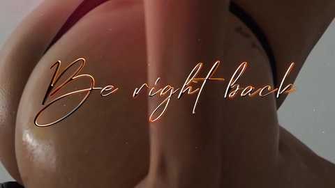 Media: Video of a close-up, glistening, brown-skinned woman's bare buttocks, with \"Be Right Back\" in elegant, gold script.