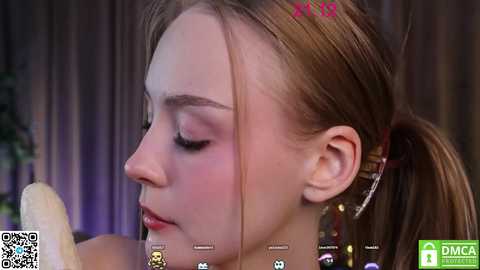 Media: A video of a young woman with light skin and light brown hair in a ponytail, wearing makeup, seen from the side, in a blurred, indoor setting.