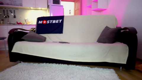 Media: A video of a cozy living room with a beige sofa covered by a white blanket, two gray throw pillows, and a \"MostBet\" flag draped on the back. The background features a modern kitchen with white cabinets, a fridge, and a pink light.