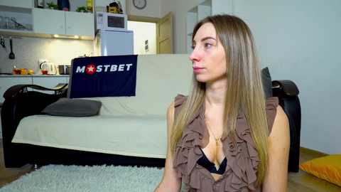 Media: Video of a light-skinned woman with long blonde hair, wearing a ruffled brown top, sitting on a soft rug in a modern kitchen-living room with white walls and black furniture.