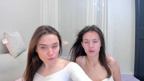 Media: Video of two young women with long dark hair, one with a white top, the other with a white bra, sitting side-by-side in a minimalist, softly lit room.