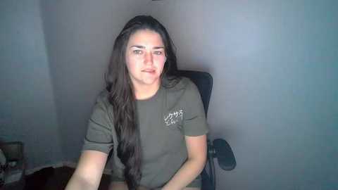 Media: Video of a young woman with long, dark hair, wearing a green t-shirt, sitting in a black chair against a plain, light grey wall.