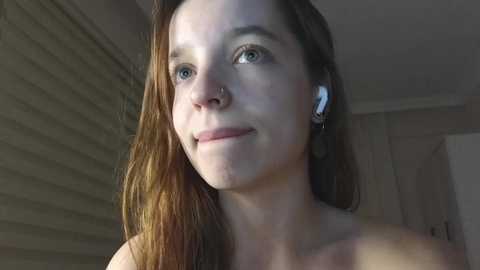 Media: Video of a fair-skinned young woman with light brown hair, wearing white earbuds, looking up thoughtfully, in a dimly lit room with beige walls.