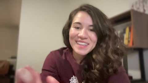 Media: Video of a smiling, light-skinned woman with wavy brown hair, wearing a maroon sweater, sitting in a cozy, dimly-lit room with bookshelves and a brown couch in the background.