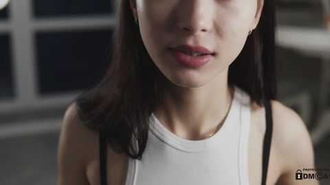 Media: Video of an Asian woman with fair skin and long black hair, wearing a white tank top. She has a neutral expression. The background is blurred, featuring a window.