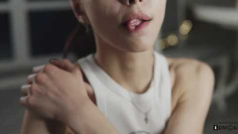 Media: A video of a young Asian woman with light skin, wearing a white tank top, hugging her arms, her lips slightly parted, in a modern indoor setting with blurred furniture.