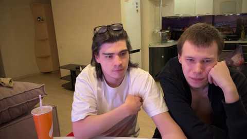 Media: Video of two young men with shoulder-length hair, wearing white T-shirts, sitting on a couch in a modern living room with a fridge, cabinets, and orange drink.