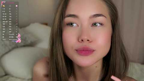 Media: Video of a young, fair-skinned woman with straight, light brown hair, green eyes, and pink lips, wearing a white top, against a blurred, beige-toned background.