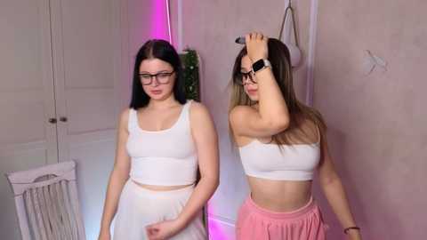 Media: Video of two young women with fair skin, one with glasses, in matching white tank tops and shorts, standing in a softly lit room with pink neon lights.