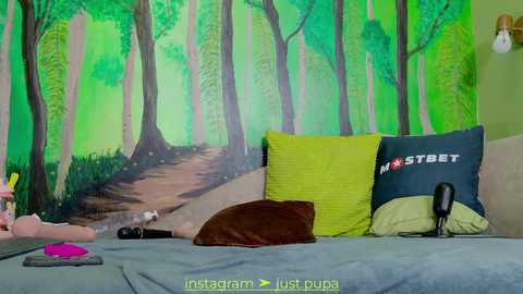 Media: Video of a cozy bedroom featuring a green forest mural, a plush brown pillow, a black pillow with \"FSTBET\" logo, and a gray blanket. A pink vibrator rests on the bed, suggesting a playful ambiance.