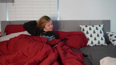 Media: Video of a young woman with blonde hair, wearing a black hoodie, lying in bed with red and gray bedding, watching TV.