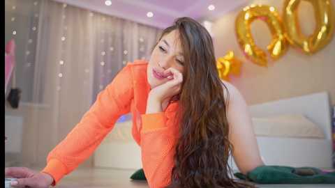 Media: Video of a young woman with long brown hair, wearing an orange sweater, leaning on a table in a modern, festive room with gold balloons and draped curtains.