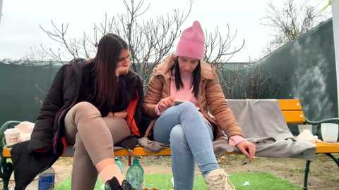 Media: Video of two women, one in a black coat, the other in a pink beanie, sitting on a bench in a winter park, reading a book.