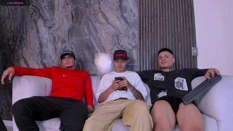 Media: Video of three young men sitting on a white couch, one wearing a red long-sleeve, another a black cap, and the third a black long-sleeve, all smoking. Background features a marble-patterned wall.