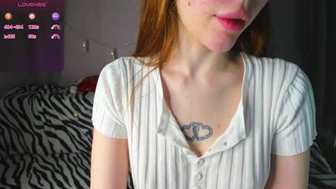 Video of a fair-skinned, red-haired young woman with a small heart tattoo on her chest, wearing a white ribbed top, against a zebra-patterned background.