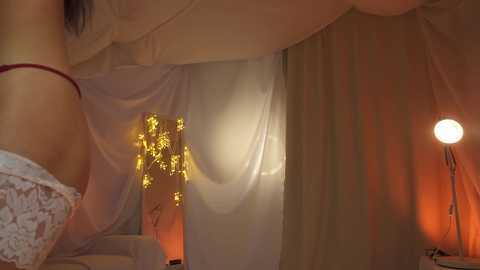 Media: A video featuring a woman in white lace lingerie, standing in a dimly lit, draped bedroom with soft, warm lighting.