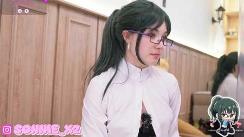 Media: Video of a young woman with long black hair in a ponytail, wearing glasses, a white shirt, and black lace bra, standing in a dimly lit room with wooden paneling.