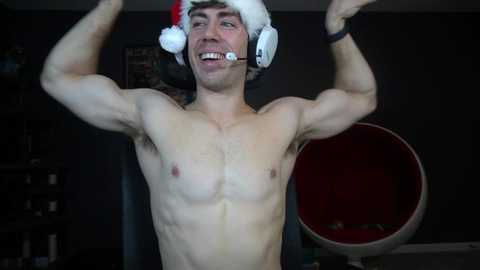 Media: Video of a muscular, shirtless man in a Santa hat with headphones, flexing his biceps. He has a white beard, and his face shows excitement. Background is a dark room with a red chair and a wall with posters.