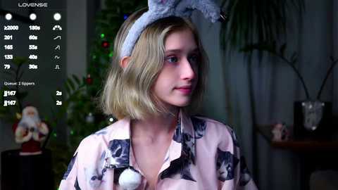 Media: A video of a young woman with shoulder-length blonde hair, wearing a light pink floral blouse and a bunny headband, seated indoors, with a festive Christmas tree and decorations in the background.