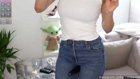 Media: Video of a person in a white t-shirt and blue jeans, holding a smartphone, in a room with a plush green toy, a TV, and a white couch.