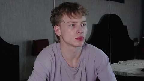 Media: Video of a young, fair-skinned boy with short, tousled brown hair, wearing a light purple shirt, sitting on a bed with a dark headboard and a white pillow, against a textured gray wall.