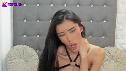 Media: Video of an Asian woman with long black hair, wearing black lingerie with a harness, lying on a tufted gray headboard. She has a soft, sensual expression.