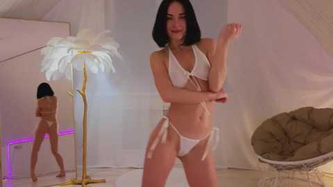 Media: Video of a slender, light-skinned woman with straight black hair, wearing a white feathered headpiece and a white bikini, dancing in a minimalist, white room with a large feathered lamp and a wicker chair.