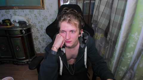 Media: Video of a young man with fair skin and short, tousled blonde hair, wearing a black hoodie and leaning on a window, looking tired and contemplative. Background features a dark wooden cabinet and floral wallpaper.