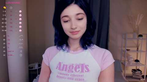 Media: Video of a young woman with long dark hair, fair skin, and wearing a pink Angels T-shirt, standing indoors with a beige wall and a bookshelf in the background.