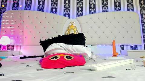 Media: Video of a plush owl, wearing a white shirt, lying on a bed with a white headboard and black-and-white striped wallpaper. The room is softly lit, with a laptop and a bright orange dildo on the bedside table.