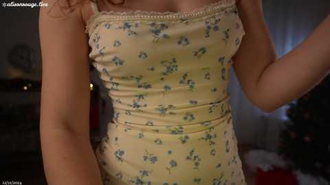 Media: A video of a light-skinned woman with curly hair, wearing a yellow floral dress with lace trim, indoors, blurred background with Christmas decorations.