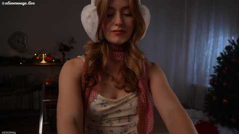 Media: Video of a young woman with fair skin and wavy blonde hair, wearing a festive outfit including a fluffy white hat and red scarf, standing indoors next to a decorated Christmas tree.