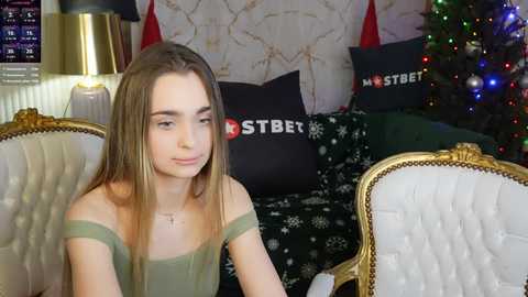 Media: Video of a young woman with long brown hair, wearing a green off-shoulder top, sitting in a luxurious, gold-framed, white tufted sofa, adorned with black and white holiday-themed pillows.