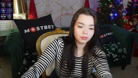 Media: Video of a young Asian woman with long, straight black hair, wearing a black-and-white striped sweater, sitting on a green couch decorated with Christmas stockings. Background includes a festive Christmas tree with colorful lights and a white wall with gold accents.