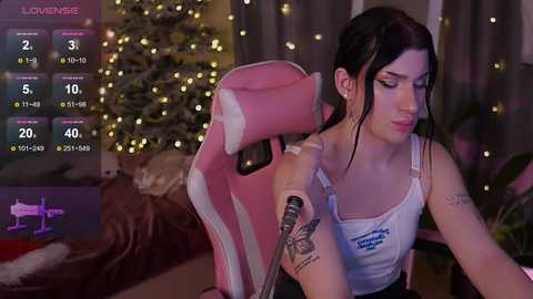 Media: A video featuring a fair-skinned, dark-haired woman in a white tank top, seated in a pink gaming chair, with a Christmas tree in the background.