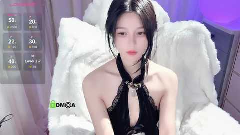 Media: Video of a young East Asian woman with pale skin, dark hair, wearing a black halterneck top, sitting on a white fur blanket in a cozy room with purple lighting.