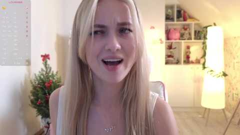 Media: A video of a blonde woman with straight hair, wearing a white top, smiling, in a brightly lit room with a Christmas tree, shelves, and a lamp.