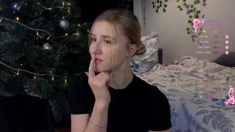 Media: Video of a blonde woman in a black shirt, resting her chin on her hand, gazing thoughtfully at a decorated Christmas tree in a cozy bedroom.