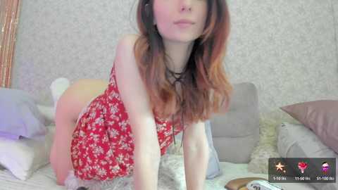 Media: Video of a slender, light-skinned woman with long, wavy brown hair, wearing a red floral dress, leaning forward on a bed. Background features patterned wallpaper, pillows, and a soft, light-colored blanket.