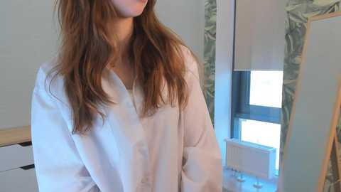 Media: Video of a woman with long, wavy brown hair, wearing a white lab coat, standing in a modern, minimalist bathroom with green marble tiles and a large window.