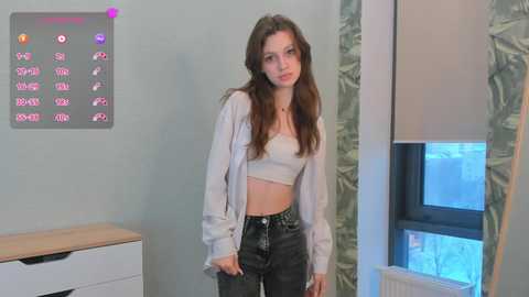 Media: Video of a young Caucasian woman with long brown hair, wearing a white crop top and high-waisted jeans, standing in a modern bedroom with a digital clock and a leaf-patterned curtain.