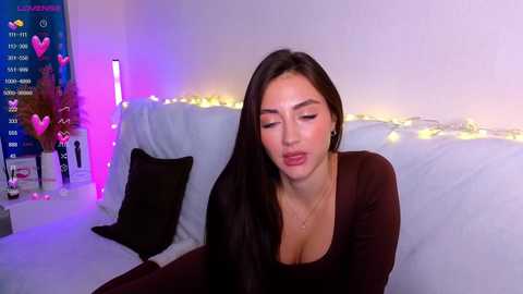 Media: Video of a Latina woman with long dark hair, wearing a low-cut brown top, sitting on a white couch in a brightly lit room with string lights and a heart-themed poster.