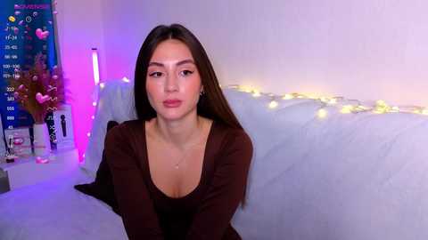 Media: Video of a young woman with long brown hair, wearing a deep V-neck brown top, sitting on a bed adorned with white bedding and fairy lights, surrounded by a colorful, festive background.