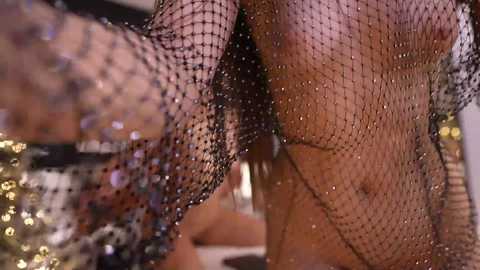 Media: Video of a woman in a black, glittery fishnet bodysuit, her arm extended forward, showcasing her bare midriff and toned physique. The background is blurred, featuring a festive, possibly indoor setting with glittering lights.