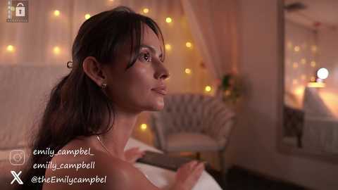 Video of a serene woman with dark hair, standing in a softly lit bedroom with string lights and a white chair.